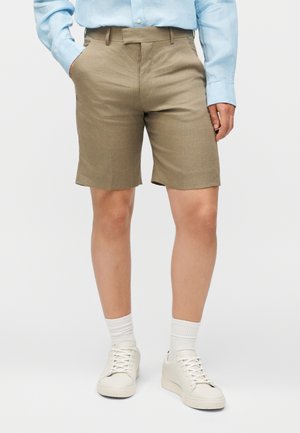 Άτομο που φοράει μπεζ σορτς μέχρι το γόνατο, ανοιχτό μπλε πουκάμισο με μακρύ μανίκι, λευκές κάλτσες και λευκά αθλητικά παπούτσια, στέκεται με το ένα χέρι στην τσέπη.