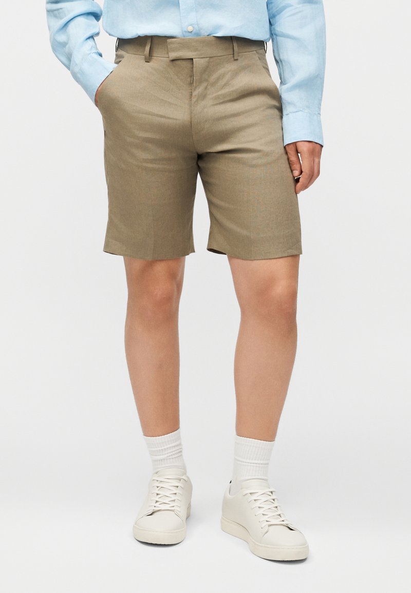 Άτομο που φοράει μπεζ σορτς μέχρι το γόνατο, ανοιχτό μπλε πουκάμισο με μακρύ μανίκι, λευκές κάλτσες και λευκά αθλητικά παπούτσια, στέκεται με το ένα χέρι στην τσέπη.