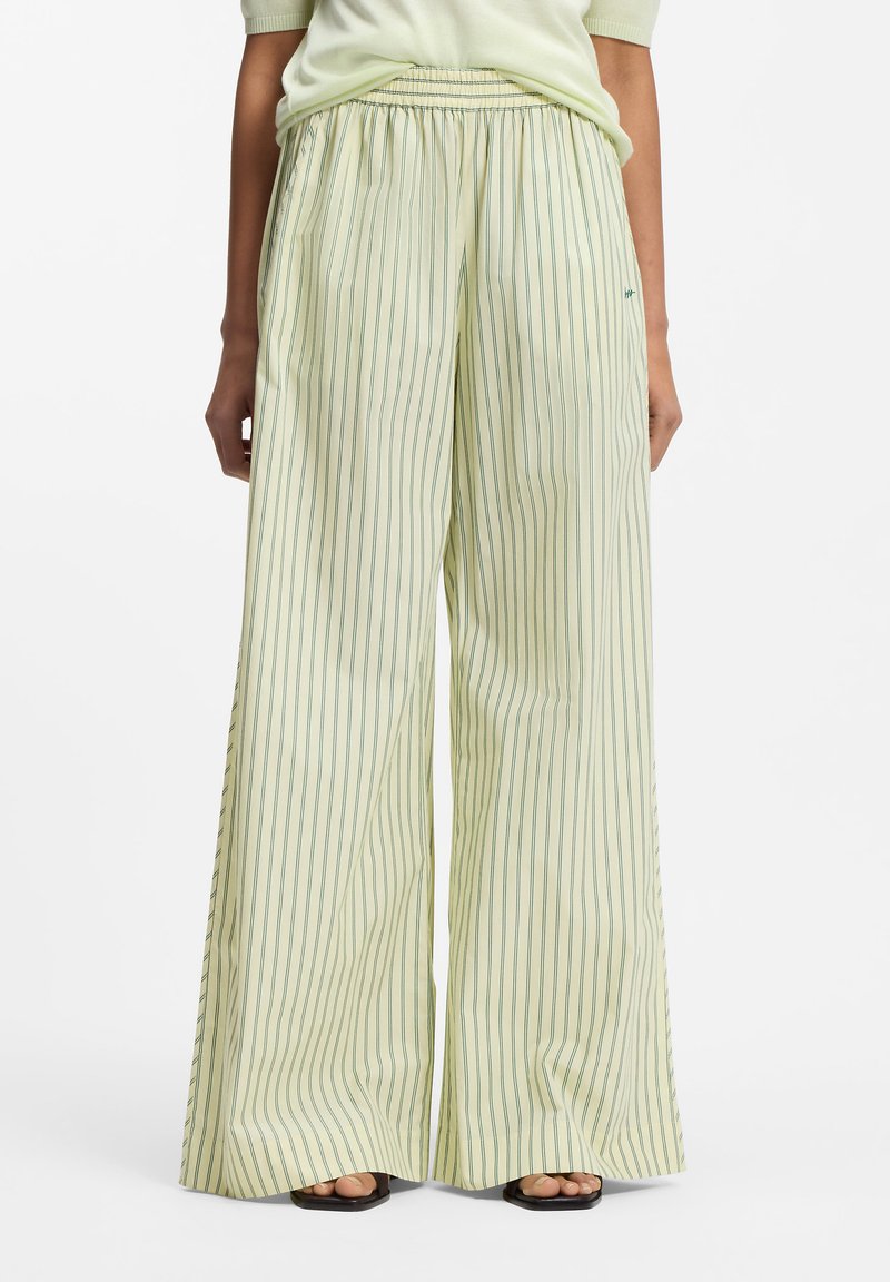 Ευρύχωροι εκρού παντελόνες με λεπτές κάθετες πράσινες ρίγες, συνδυασμένοι με ανοιχτό πράσινο τοπ και μαύρα σανδάλια με ανοιχτά δάχτυλα.