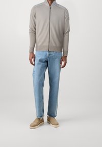 Světle šedý svetr na zip s ribovanou texturou, kontrastními detaily na ramenou, kombinovaný se světle modrými džínami rovného střihu a béžovými nazouvacími botami.
