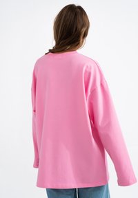 Oversize t-shirt z długim rękawem w jaskrawym różowym kolorze, wykonany z bawełny, z okrągłym dekoltem i widocznymi detalami szycia. Lekko zaokrąglony dół.