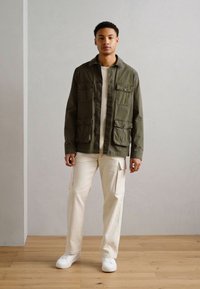 Chaqueta verde oliva con cuatro bolsillos frontales, camisa color crema y pantalones de corduroy beige claro. Zapatillas blancas completan el look.