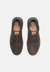 Zapatos de cuero marrón oscuro con punta redonda, cordones grises y plantillas color beige con un logo. Textura suave con un diseño minimalista.