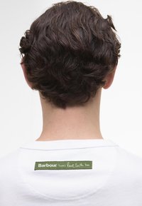 Camisa branca de manga curta com um rótulo verde e texto branco nas costas, que diz "Barbour também ama Paul Smith." Tecido texturizado visível.