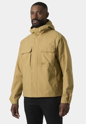 Mężczyzna w beżowej kurtce z kapturem, z dwoma kieszeniami na piersi i czarnych spodniach, stojący na tle jasnego, jednolitego tła.