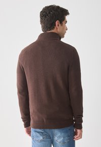 Maglione lavorato a maglia marrone con un alto collo e accenti texturizzati sulle spalle, abbinato a jeans blu. Il tessuto appare morbido e caldo.