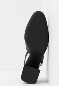 Semelle de chaussure habillée en cuir noir tournée vers le haut, positionnée contre un coin de mur et de sol blancs, mettant en valeur la forme et la texture de la chaussure.