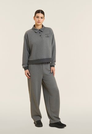 Donna in piedi che indossa una felpa grigia a maniche lunghe con colletto e pantaloni grigi larghi con scarpe nere su uno sfondo semplice.