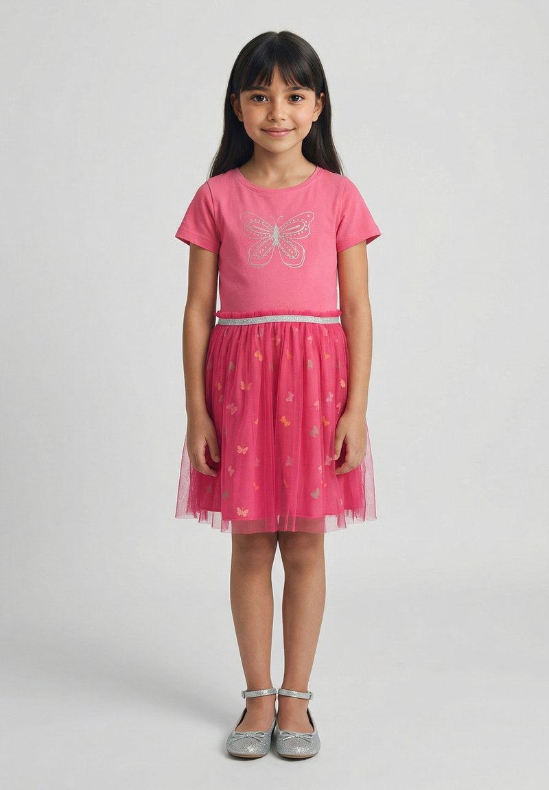 Ragazza giovane con lunghi capelli scuri, indossa un vestito t-shirt rosa con farfalle e scarpe argentate, in piedi davanti a uno sfondo neutro.