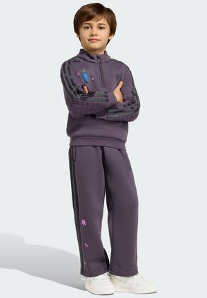 Junger Junge steht mit verschränkten Armen, trägt einen lila Trainingsanzug mit schwarzen Streifen und weiße Sneakers vor einem einfarbigen Hintergrund.