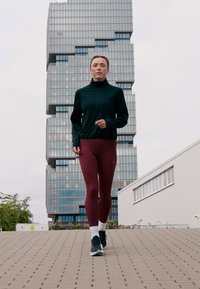 Kvinde i sort jakke og bordeaux leggings, der jogger på en asfalteret sti med en moderne glastbygning i baggrunden.