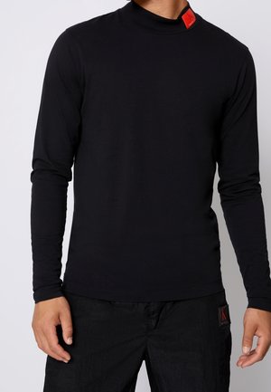Mand iført en tætsiddende sort langærmet skjorte med røde Calvin Klein logo patches på kraven og bukselommen.