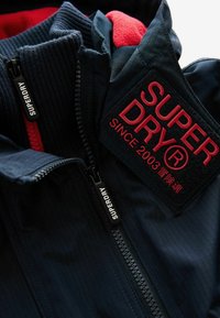 Giacca blu marina con interno in pile rosso, tessuto texturizzato e tirazip neri. Presenta una toppa con "SUPERDRY" e dettagli del marchio.