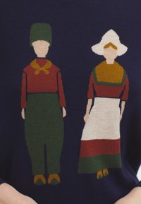 Maglione lavorato a maglia con sfondo navy che presenta due figure stilizzate. La figura a sinistra indossa pantaloni verdi e una maglietta rossa; quella a destra indossa un abito bianco.