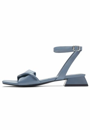 Sandales plates en cuir bleu clair avec une sangle nouée torsadée et une boucle à la cheville. Dotées d'un petit talon angulaire. Texture lisse.