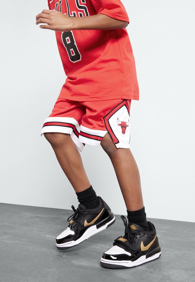 Czerwona koszulka Chicago Bulls z czarnymi i białymi detalami; czerwone szorty z logo drużyny i kontrastowymi białymi wykończeniami; czarne sneakersy ze złotymi akcentami.