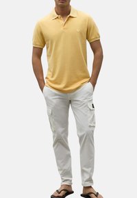 Camisa polo amarilla, pantalones cargo blancos con bolsillos laterales y chanclas negras. Conjunto informal con un ajuste relajado y suave textura.