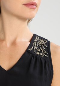 Musta mekko, jossa on teksturoitu kangas, V-pääntie ja koristeellinen helmillä suunniteltu kukkakuvio vasemmassa olkapäässä.
