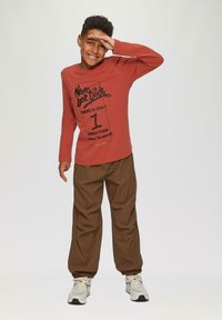 Lächelnder Junge posiert mit der Hand, die die Augen schützt, trägt ein rostfarbenes Langarmhemd mit motivierendem Text, braune Hosen und weiße Sneaker vor weißem Hintergrund.