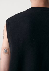 Črn brezrokavnik s pletenim izrezom in teksturo; delno viden tatu sonca na roki.