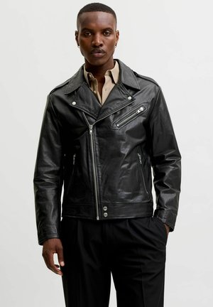 Modèle masculin portant une veste en cuir noir de motard avec des poches zippées et une chemise beige sur un fond clair uni.