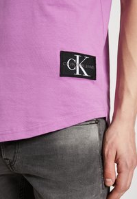 Detailní záběr osoby ve růžovém tričku Calvin Klein Jeans a vybledlých šedých džínách, na tričku je vidět nášivka s logem CK.