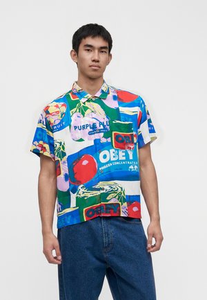 Young man wearing a short-sleeve button-up shirt with colorful fruit and text prints, paired with loose blue jeans, standing against white background.