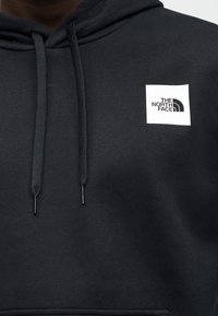 Gros plan d'un sweat à capuche noir avec des cordons et un logo carré blanc indiquant "The North Face" sur la poitrine.