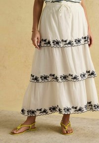 Biela viacvrstvová dlhá sukňa so čiernym kvetinovým výšivkou pozdĺž lemu. Materiál pôsobí ľahko a vzdušne. Nosená so žltými sandálmi.