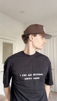 Černé tričko s přiléhavým oversized střihem, na kterém je bíle natištěný text "Mám rád špatná rozhodnutí, promiň mami." Kombinováno s hnědou baseballovou čepicí.