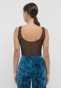 Una donna con capelli scuri raccolti in un basso chignon indossa una maglietta senza maniche in mesh nera e pantaloni in velluto a coste blu, mostrando tatuaggi su entrambe le braccia superiori.