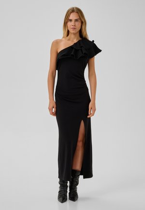 Femme portant une robe noire asymétrique avec un détail volanté et une fente à la cuisse, associée à des bottes noires pointues, debout sur un fond gris.