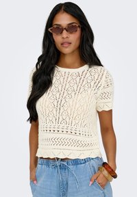 Pull crème à manches courtes en maille avec motif ajouré ouvert et ourlet festonné, associé à un short en denim. Le mannequin porte des lunettes de soleil marron à monture œil de chat.