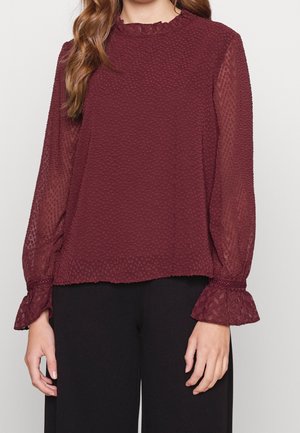 Bordowa bluzka z teksturowanym przodem, przezroczystymi długimi rękawami i marszczonym dekoltem. Posiada ściągacze przy rękawach i luźny krój. Połączona z czarnymi spodniami.