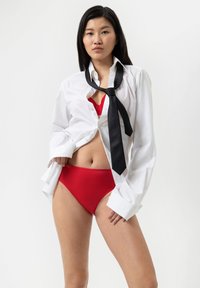 Witte shirt gedeeltelijk geknopt met opgerolde mouwen, zwarte stropdas losjes geknoopt en rode bikini onderkant. Model staat tegen een neutrale achtergrond.