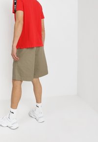 T-shirt rosso a maniche corte con accenti neri ai lati, abbinato a pantaloni corti khaki di lunghezza al ginocchio. Sneakers bianche e calze di lunghezza alla caviglia completano l'outfit.