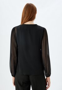 Blusa nera con maniche a sbuffo trasparenti, scollatura rotonda e retro liscio. Il tessuto si drappeggia dolcemente, creando una silhouette fluida.