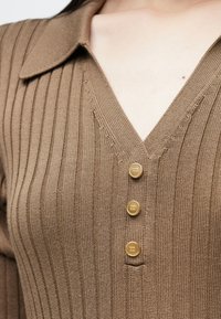 Καφέ πλεκτό τοπ με ραβδώσεις, με V-λαιμόκοψη, γιακά και τρία χρυσαφί κουμπιά μπροστά. Λεπτή υφή και διακριτικές λεπτομέρειες ραφής.