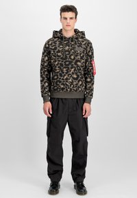 Modello maschile che indossa un hoodie a stampa leopardata verde scuro e marrone con pantaloni cargo neri e stivali neri, in piedi davanti a uno sfondo chiaro e semplice.