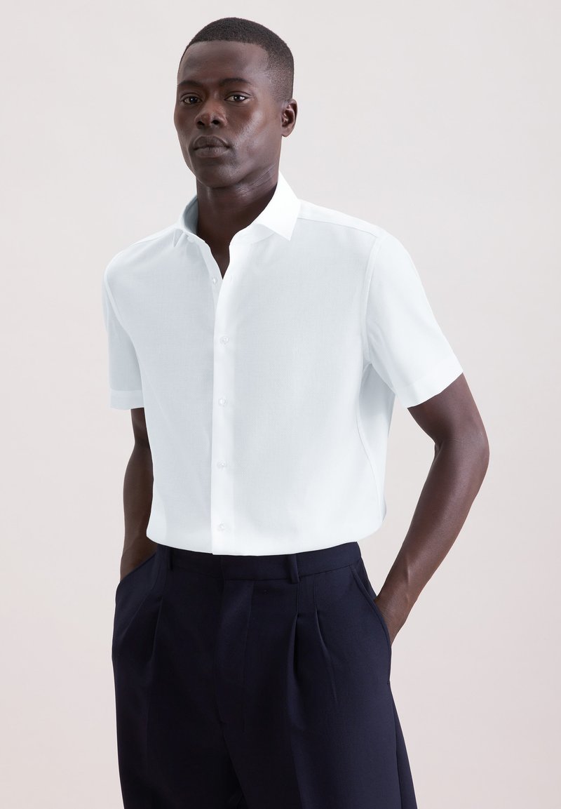 Homme portant une chemise blanche à manches courtes boutonnée, rentrée dans un pantalon à plis bleu marine foncé taille haute, les mains dans les poches.