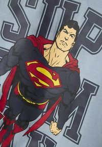 Superman graphic t-shirt met een blauwe achtergrond, vet zwart tekst en een illustratie van Superman met rode en gele accenten.