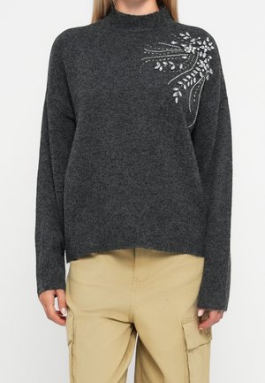 Pull en maille gris foncé avec ornements décoratifs en feuille d'argent et perles sur l'épaule gauche supérieure, porté avec un pantalon cargo beige.