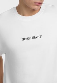 Valge puuvillane T-särk, millel on must tikandiga "GUESS JEANS" logo, mis asub ees keskel. Lühikesed varrukad ja ümmargune kaelus. Sile tekstuur.