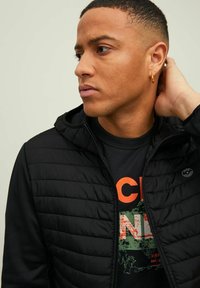 Schwarze gesteppten Jacke mit Kapuze, Reißverschluss und rundem Logo. Getragen über einem schwarzen Shirt mit orangefarbener und grüner Schrift sowie Grafiken.