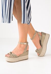 Gambe femminili che indossano sandali con zeppa beige e cinturini alla caviglia, abbinati a una gonna a righe blu e bianche.