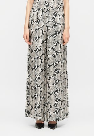 Γυναίκα που φοράει παντελόνι φαρδύ με ασπρόμαυρο μοτίβο φίδι και μαύρα μυτερά τακούνια, σε λευκό φόντο.