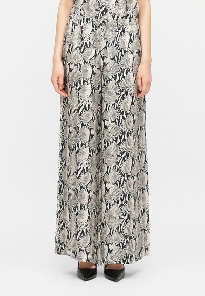 Femme portant un pantalon à jambes larges avec un motif peau de serpent noir et blanc et des talons pointus noirs sur fond blanc.