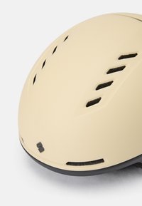 Beżowy kask rowerowy z wentylacją i gładką matową powierzchnią, z kontrastową czarną obręczą i detalem logo na dole.