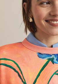 Trui met een bloemmotief in oranje en blauwe tinten, met een geribde halslijn. Blauwe overhemd met kraag zichtbaar eronder. Gouden oorstekers.