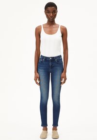 Débardeur blanc à c ôtes, jeans skinny bleu avec un léger délavage et chaussures beige à enfiler. Le modèle se tient devant un fond blanc uni.